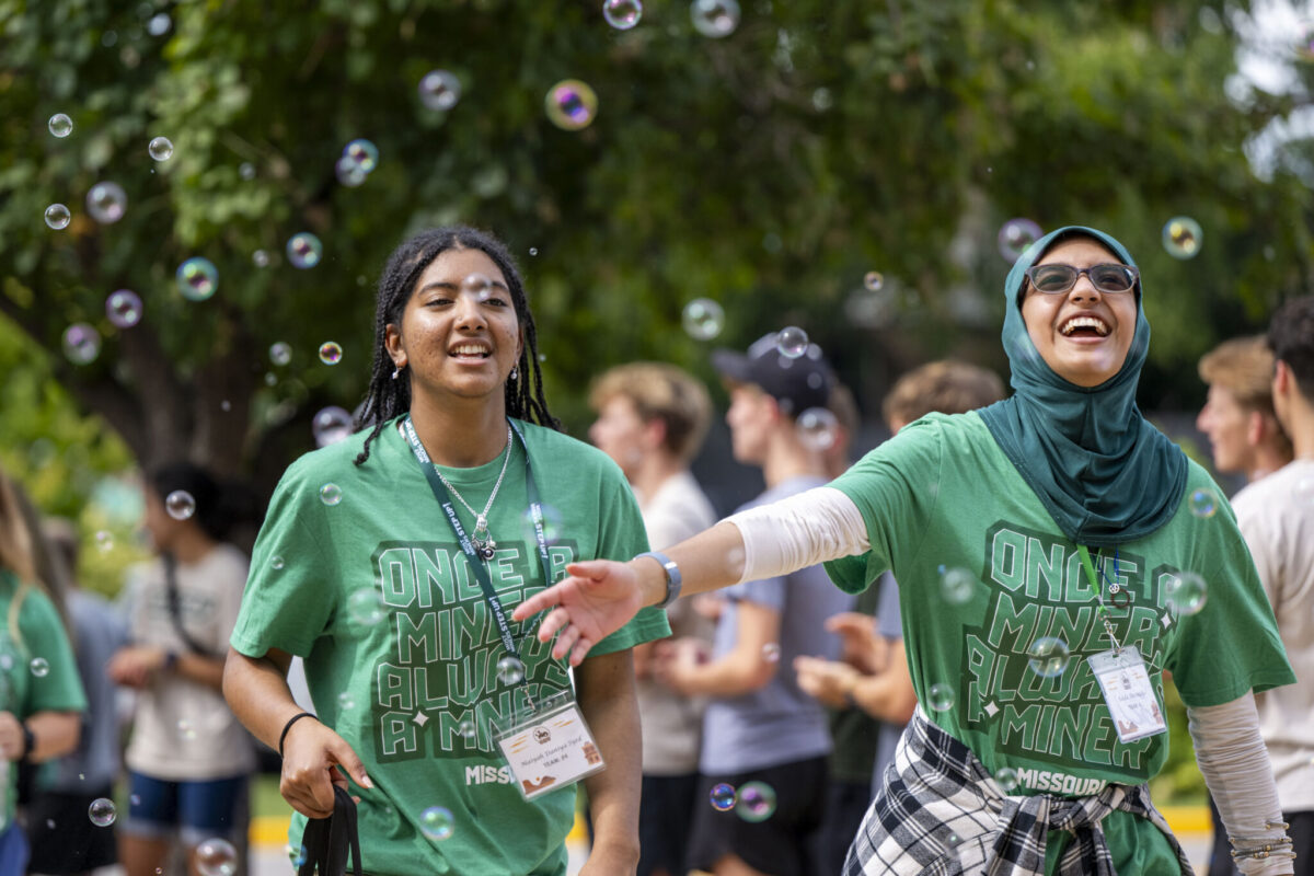 Students in green Missouri S&T shirts walk outdoors toward New Student Convocation as bubbles float through the air and other students gather in the background.