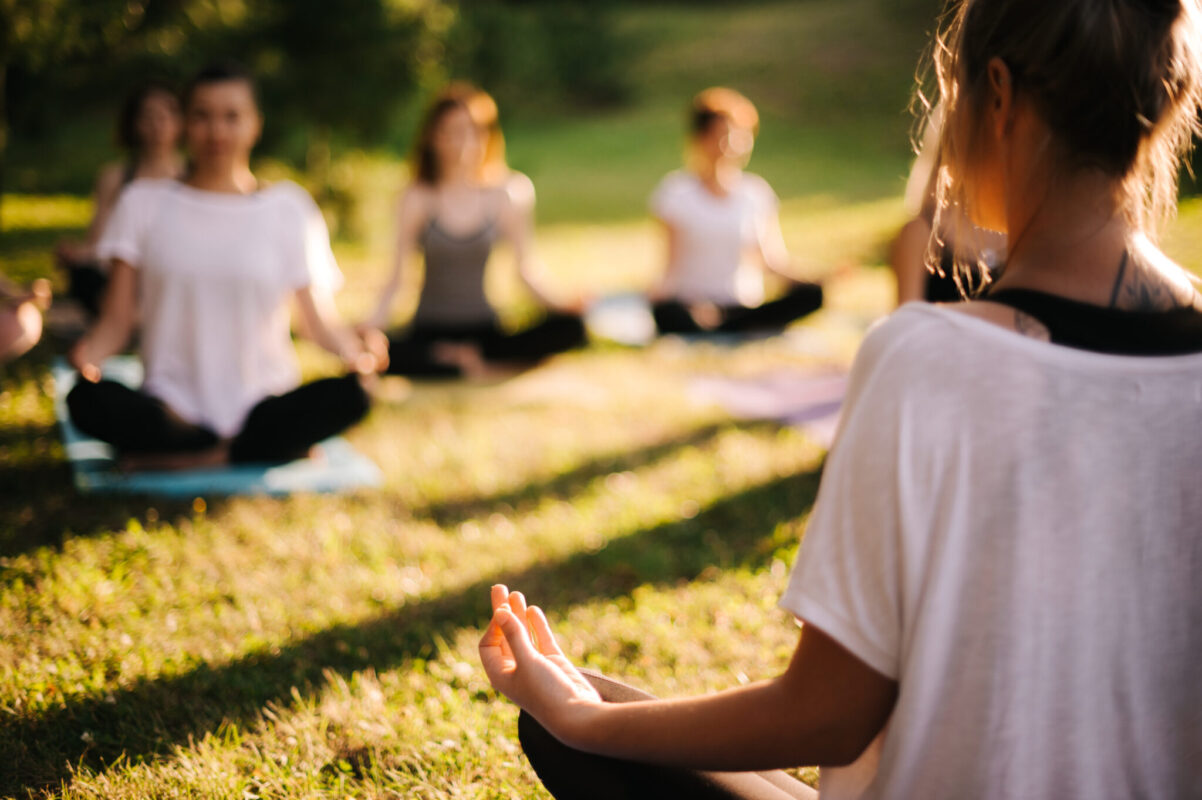 Group of young sporty woman practicing yoga lesson with instructor, sitting in lotus pose. Focus of camera at teacher rear view. Photo from back of instructor. Group of meditating people in background.
