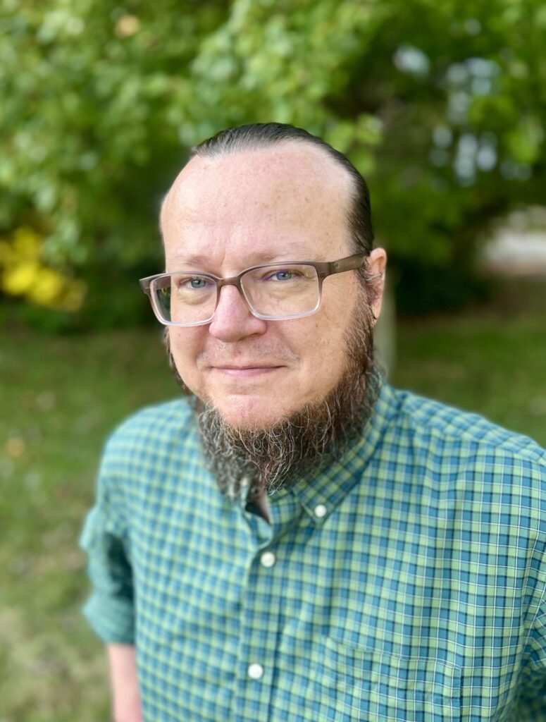 Portrait of a person wearing glasses and a green-and-blue plaid shirt, standing outdoors with leafy trees and grass softly blurred in the background.