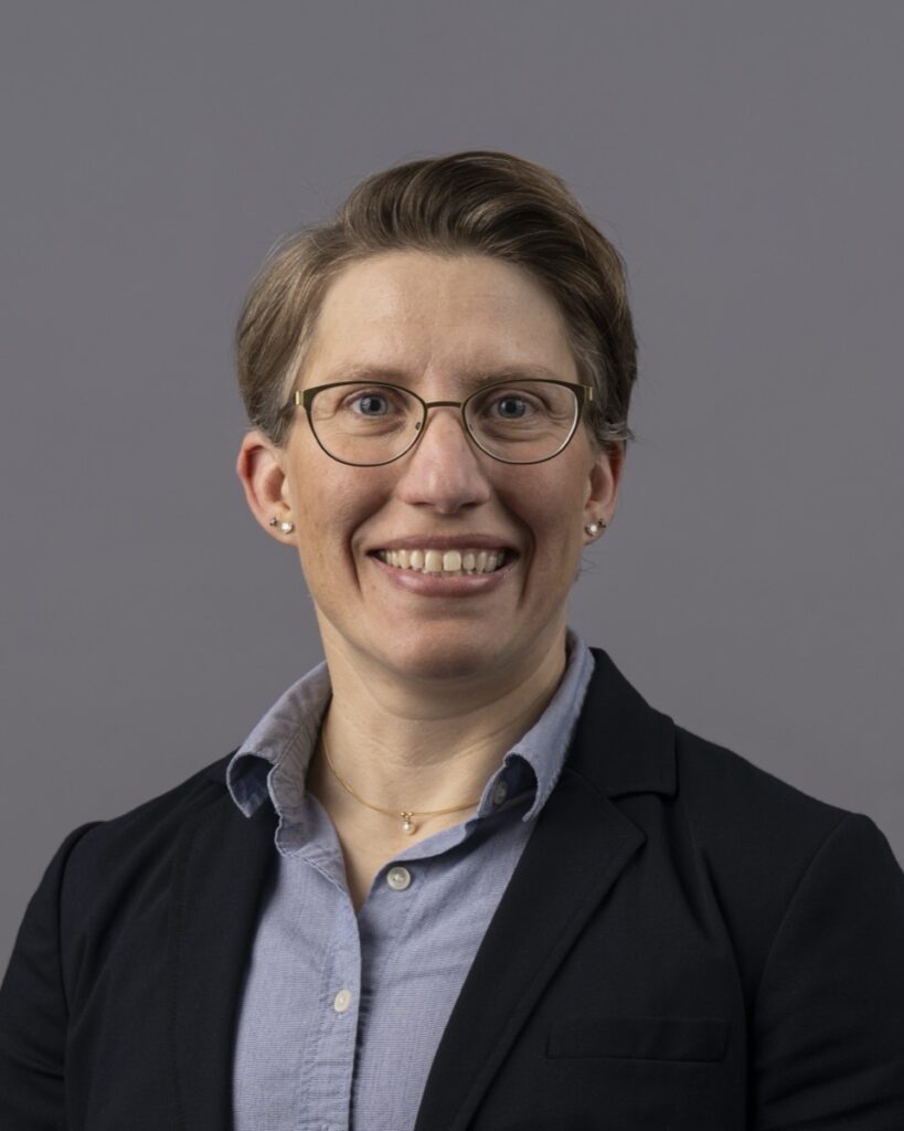 Portrait of a person wearing glasses, a dark blazer and a light blue collared shirt against a plain gray background.