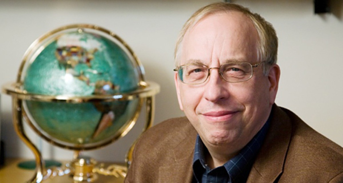Portrait of a person wearing glasses and a brown jacket, seated beside a decorative green globe.