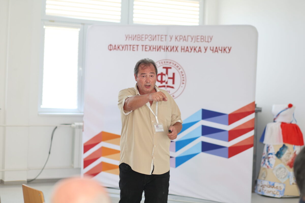 A person speaks into a microphone and gestures during a presentation in front of a university-branded backdrop with colorful geometric designs.