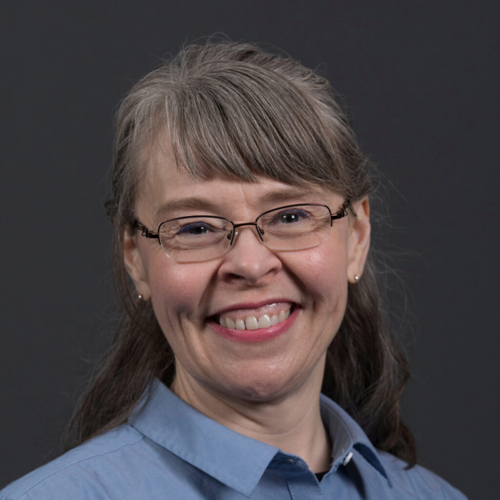 Professional headshot of a person wearing glasses and a light blue collared shirt, facing the camera against a dark gray background. The person has medium-length hair with bangs and is smiling.