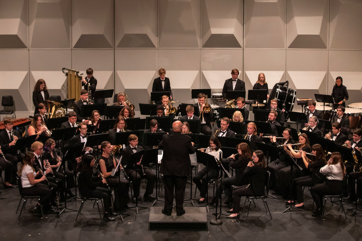 Large concert band performing onstage with a conductor, featuring woodwind, brass and percussion musicians.