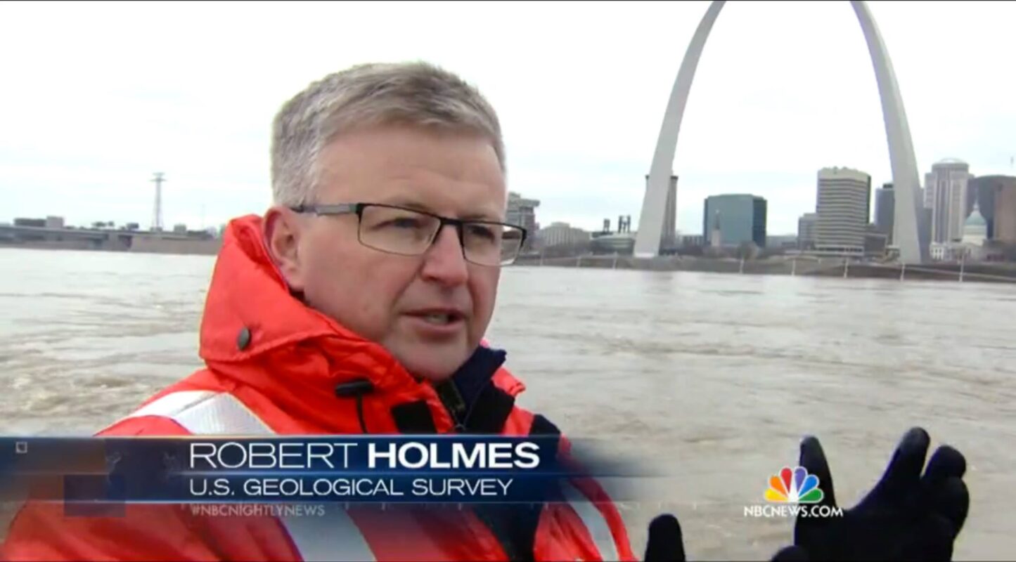Television news screenshot showing Robert Holmes wearing a bright orange jacket and black gloves near a river. The St. Louis skyline and the Gateway Arch are visible in the background. A lower-third graphic identifies Holmes as "Robert Holmes, U.S. Geological Survey," and the NBC News logo appears in the corner.