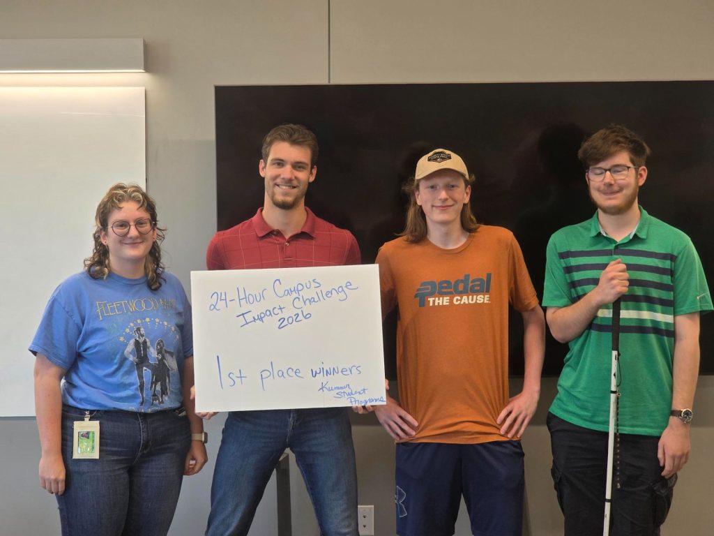 Four people stand together in a classroom, with one holding a whiteboard that reads, “24-Hour Campus Impact Challenge 2026, 1st place winners.”